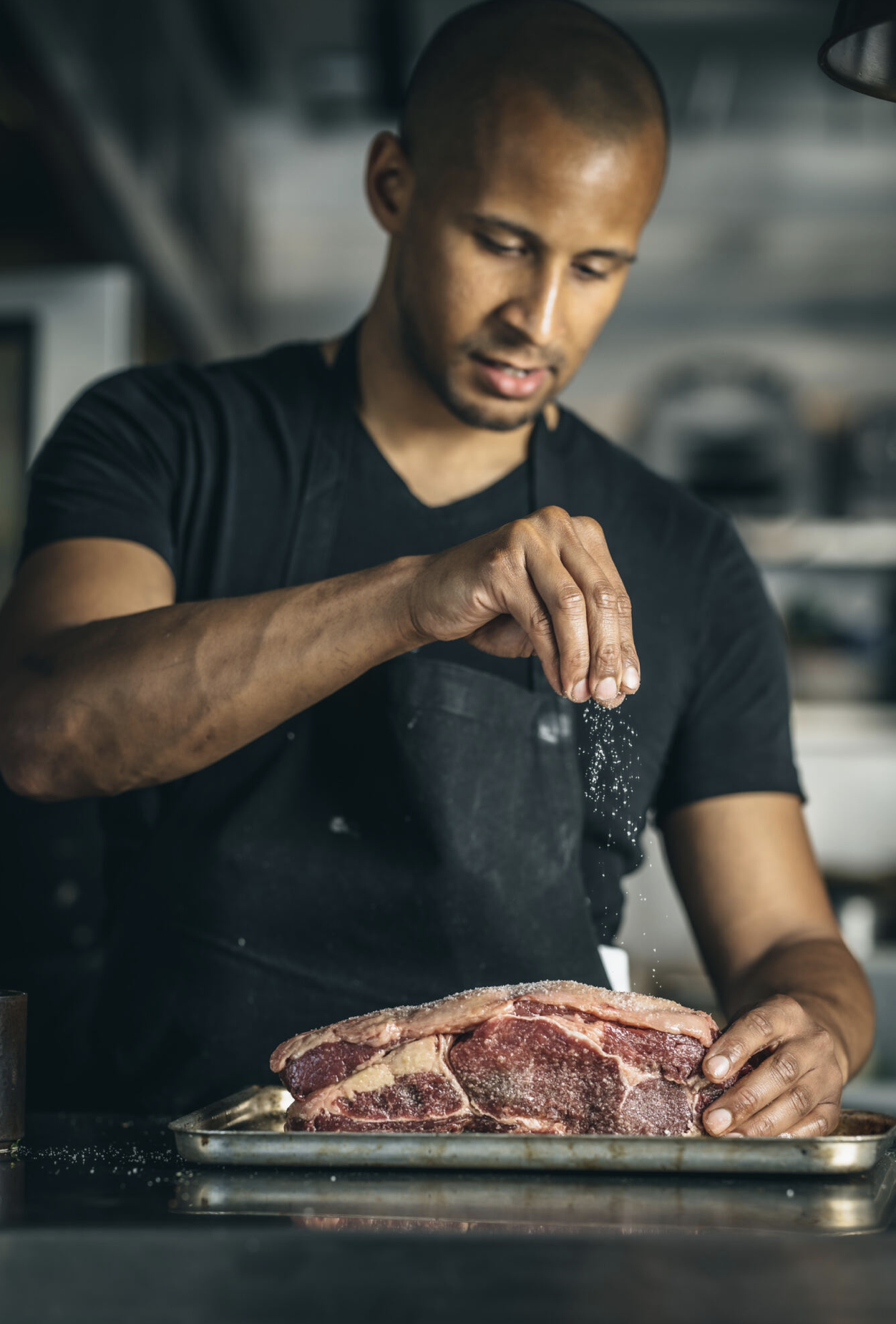 Chef Remi Williams seasoning a large cut of beef in the kitchen at Smoke & Salt in Tooting Broadway.